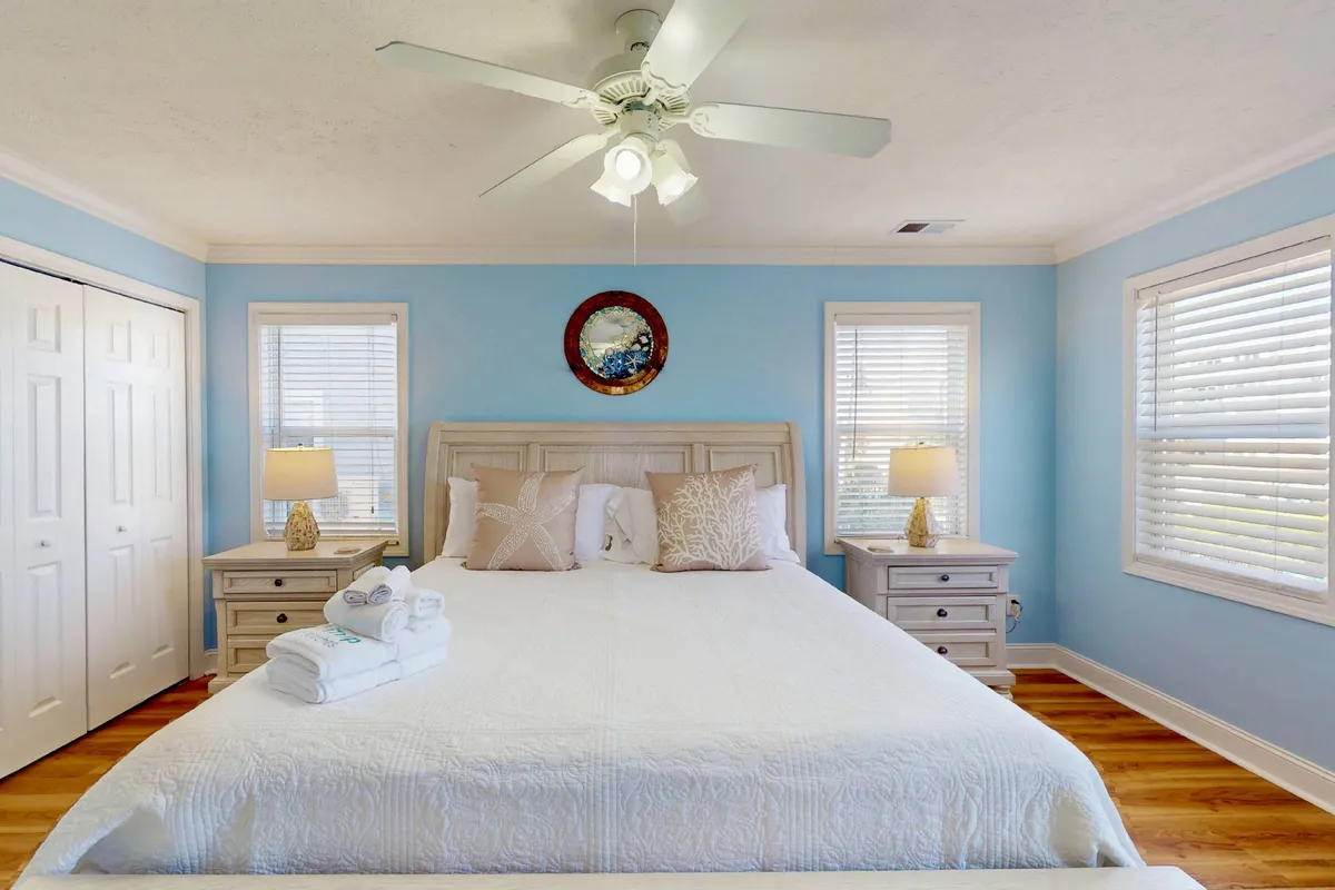 A tranquil bedroom with a plush, well-dressed bed, decorative pillows, nightstands with lamps, and a wall mirror. The room is filled with soft, natural light, creating a peaceful atmosphere ideal for restful nights.