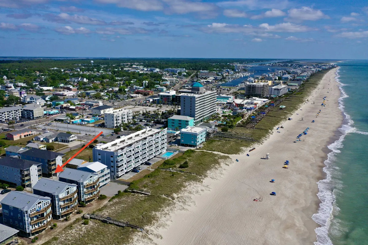 A panoramic view of beachfront properties standing right along a broad, sandy beach with the ocean waves gently rolling in. The townscape is visible beyond, suggesting nearby shops and restaurants, while people can be seen engaging in various beach activities.