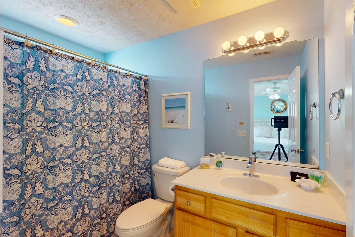 A fresh, modern bathroom with a blue patterned shower curtain, bright vanity with a sink, wall mirror, and framed artwork. The space feels airy and newly updated, signaling a clean, inviting place for morning and evening routines.