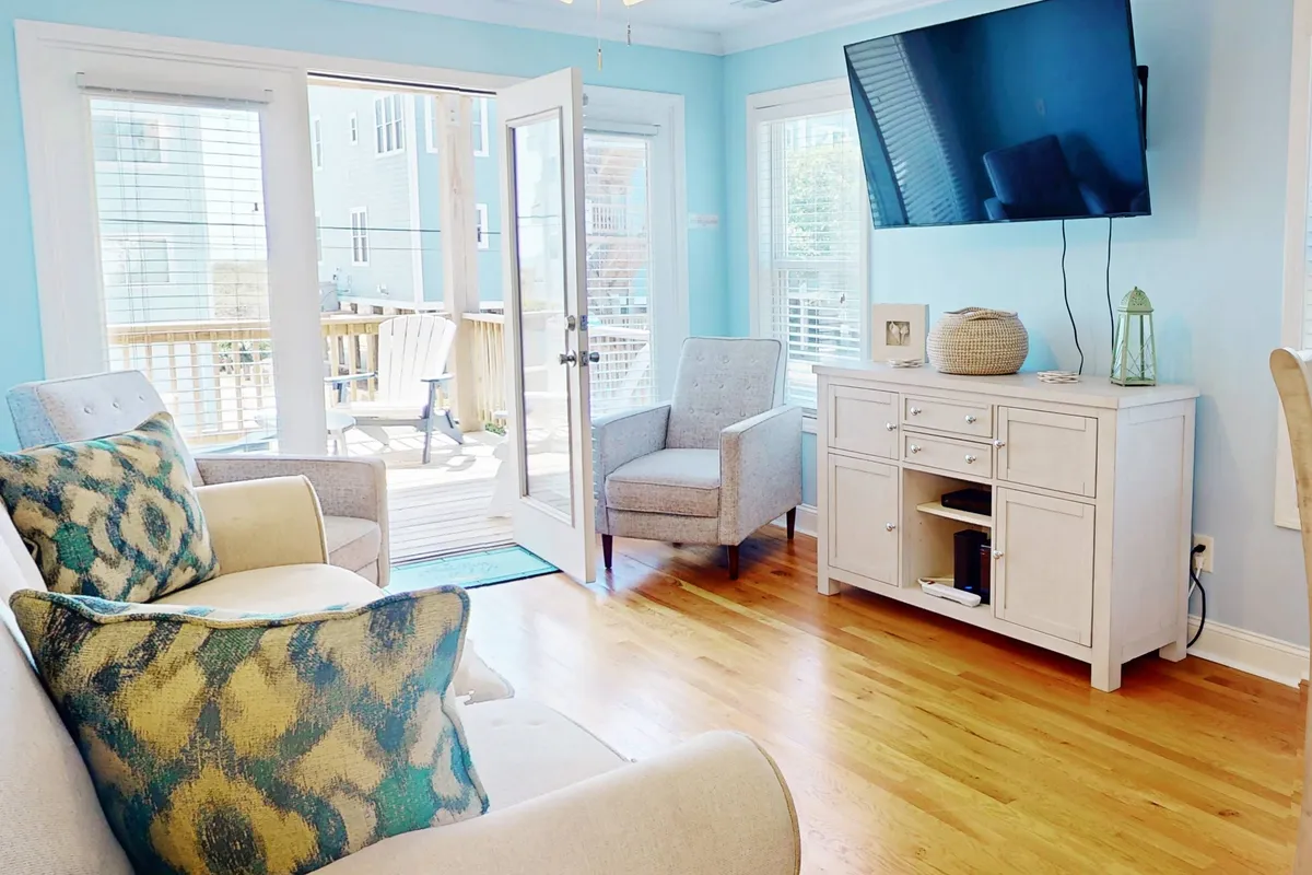 A comfortable living room setup with a sofa featuring bold patterned cushions, light wood furniture, and a TV mounted on the wall. Open doors lead out to a deck, creating a seamless connection between indoor comfort and outdoor relaxation.