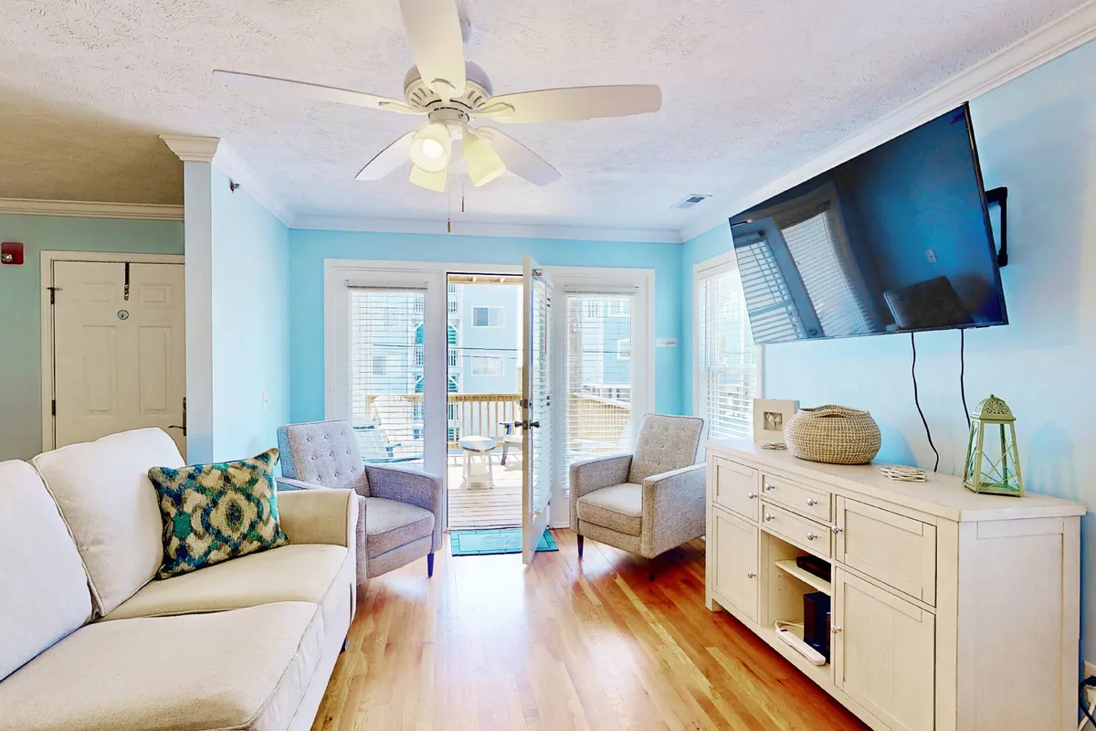 An interior view of a cozy living area with a sofa, two armchairs, and a television. The open doors lead seamlessly to the deck, inviting natural light and providing a glimpse of the outdoor space beyond. The décor is warm and inviting, emphasizing the flow from indoors to outdoors.