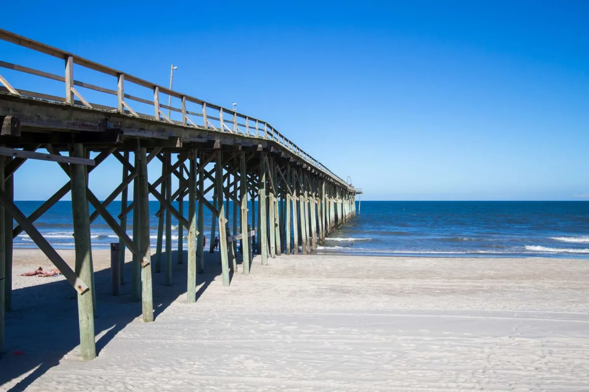 A wide wooden pier stretches out over the gentle ocean waves, with sandy beach in the foreground and the open sea beyond. The scene feels peaceful yet active, hinting at opportunities for fishing, walking, or simply enjoying the coastal views.