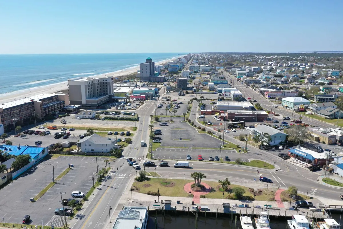 A sweeping view of the ocean coastline with the city skyline in the background, main roadways, parking lots, and commercial buildings bustling with activity. The image captures the energetic atmosphere of Carolina Beach, with visible areas for beach activities, shopping, dining, and sightseeing. The golden light of sunset infuses the scene with warmth and vibrancy.