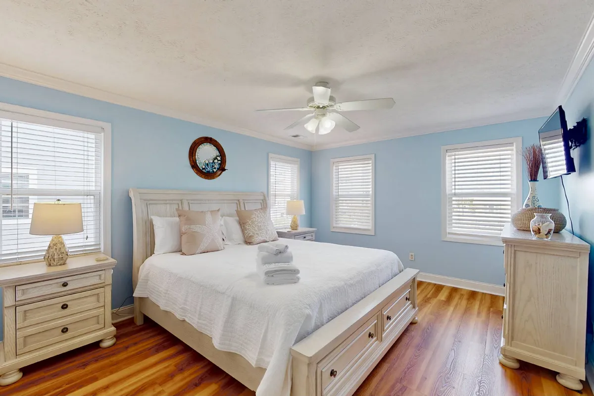 A serene bedroom with a comfortable bed, nightstands, windows letting in natural light, tasteful wall decor, and a television. The space exudes calm and offers a peaceful spot for winding down, ideal for a queen bedroom escape.