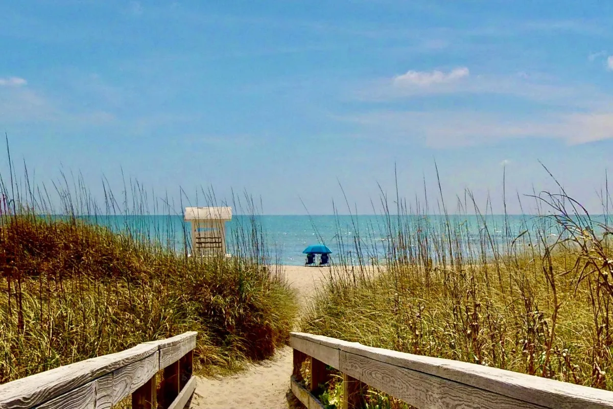 A wooden walkway leads directly to a wide, sandy beach with the ocean stretching out in the background. A blue umbrella and a lifeguard stand are visible, as are gentle waves rolling in. The scene captures the ease of beach access, family-friendly amenities, and the welcoming, tranquil mood of the shore.
