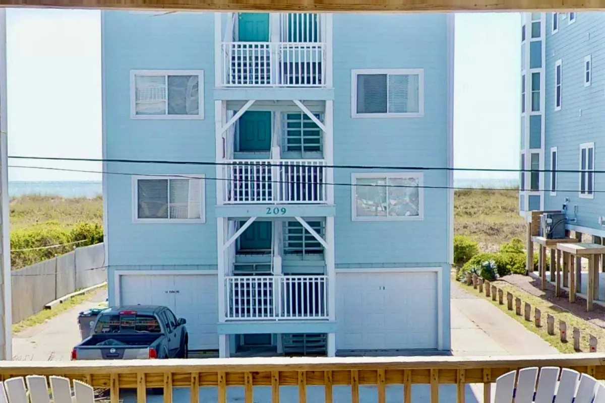 An exterior angle shows the building's blue facade, white balconies, and a parking area with a partial view of the ocean in the background. The setup communicates the proximity to the beach and the deck's elevated nature above the carport, supporting the theme of privacy and sweeping views.