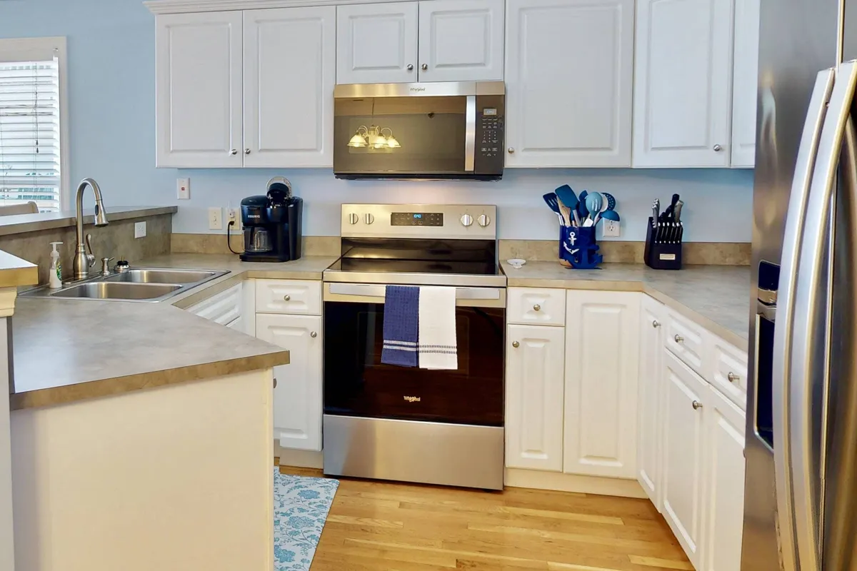 Bright, stylish kitchen with sparkling stainless steel appliances, crisp white cabinetry, blue accent details, and warm wooden flooring. The kitchen is ready for group meal prep or casual breakfasts, with plenty of space for multiple people to gather and create.