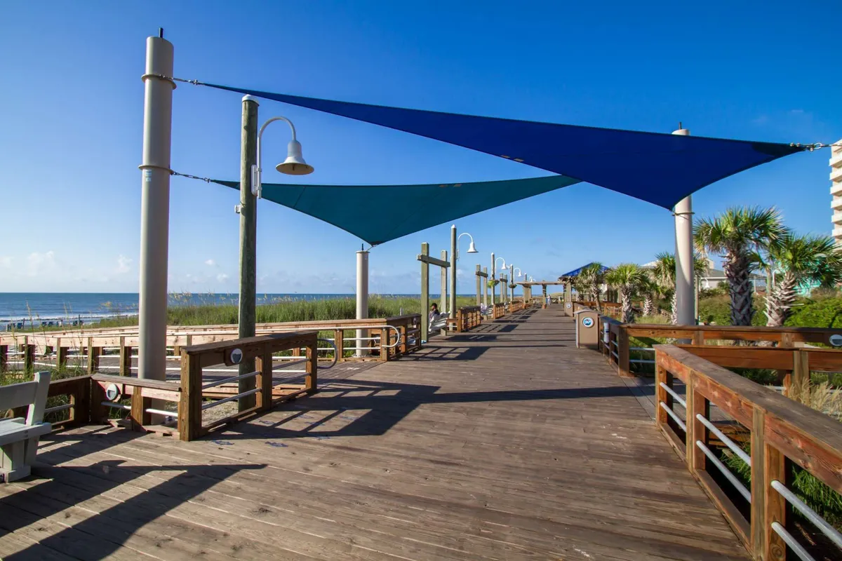 A tranquil boardwalk scene with blue shade sails, lush greenery, and views extending toward the distant beach and ocean. The pathway encourages walking, biking, and relaxing outdoors, suggesting easy access to nature and recreational activities.
