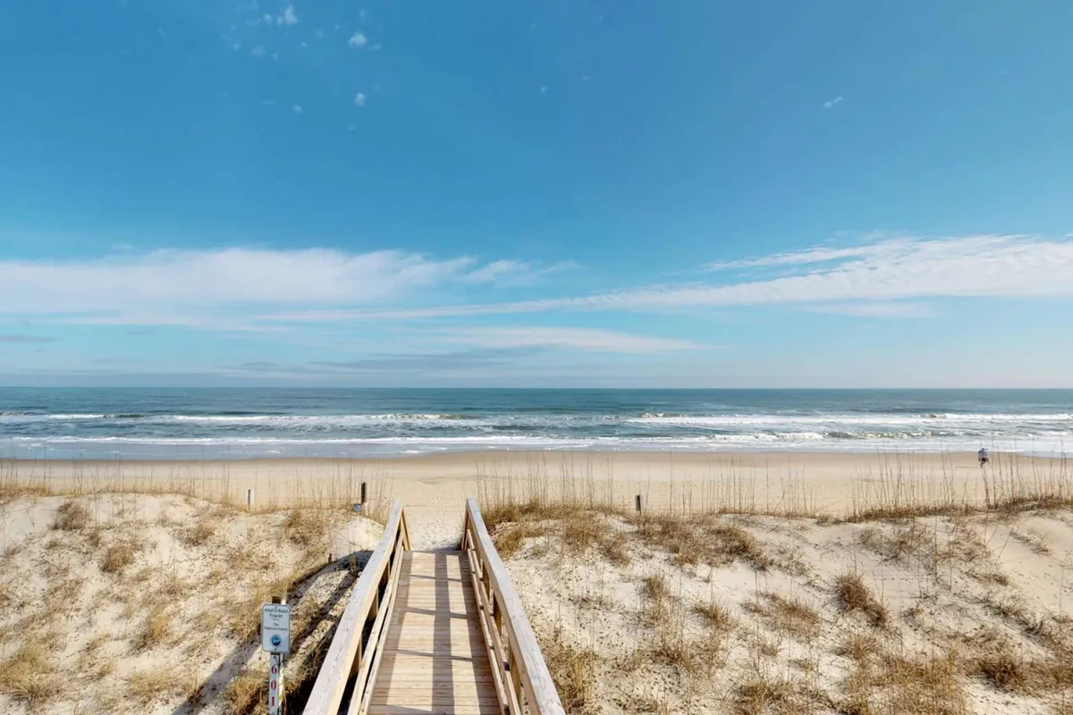 A scenic boardwalk framed by sand dunes leads out to the ocean. The early morning or late afternoon light creates a relaxed atmosphere, hinting at family activities like walks with coffee, building sandcastles, or sunbathing. The sand and waves invite visitors to explore the shoreline.