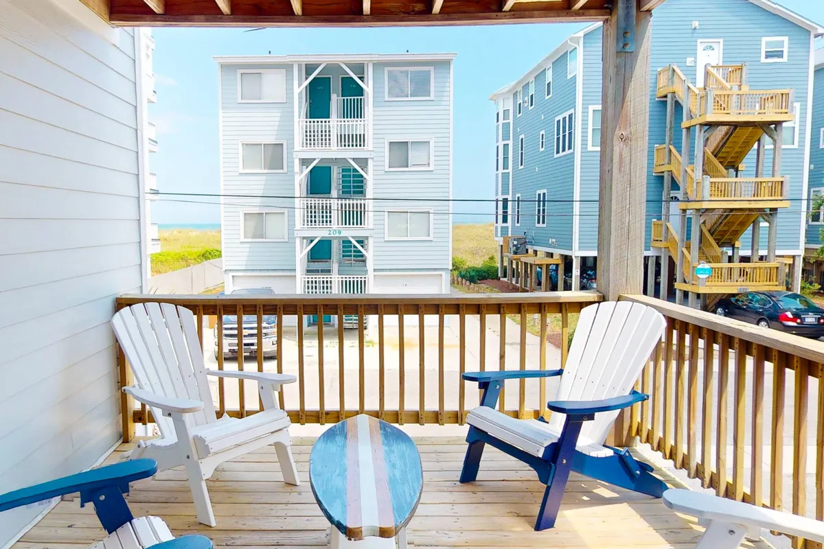 The image showcases a breezy condo balcony furnished with inviting Adirondack chairs and a decorative table, overlooking a beautiful beach view across the street. The wooden railing frames the scene, offering a perfect spot to relax, socialize with friends, and soak in the coastal ambiance at sunrise or during an evening gathering.