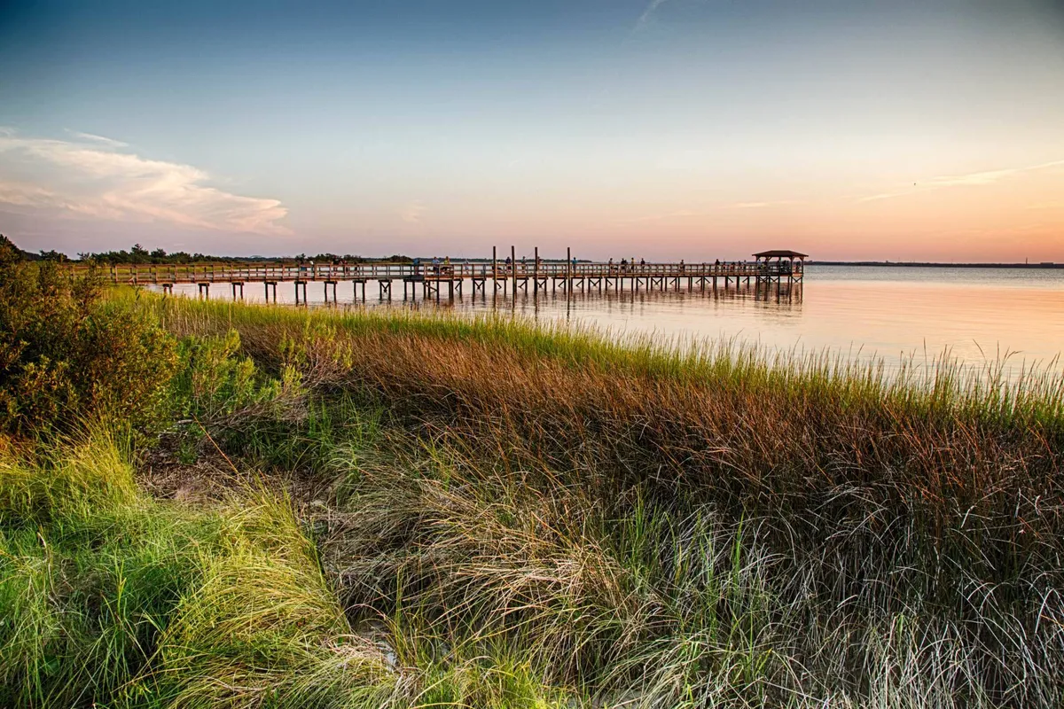 A serene sunset view over calm water, with a wooden dock extending out and warm evening light reflecting on the surface. The scene evokes peaceful moments spent by the lake, such as paddle boating or simply unwinding at dusk.