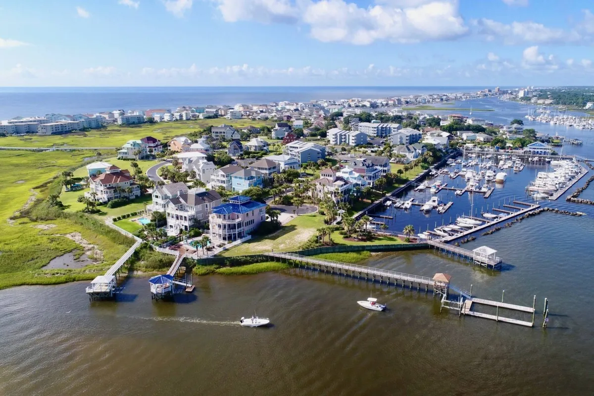 A lively marina filled with boats is bordered by colorful houses, green marshland, and the deep blue of the ocean. The atmosphere suggests a hub of water-based activities, from boating and fishing to leisurely walks along the docks.